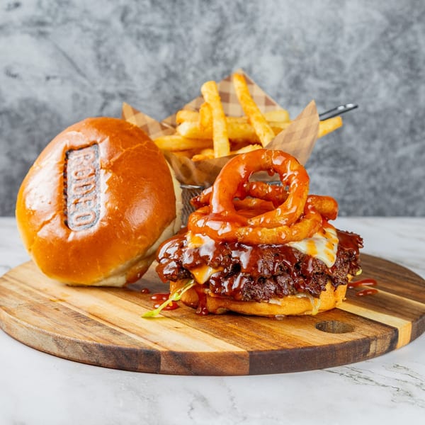 Rodeo burger with BBQ sauce, onion rings, and fries on a wooden board.