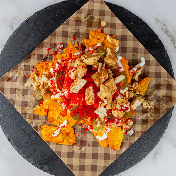 Chicken nachos with red spice, jalapeños, and white sauce on checkered paper and a dark plate.