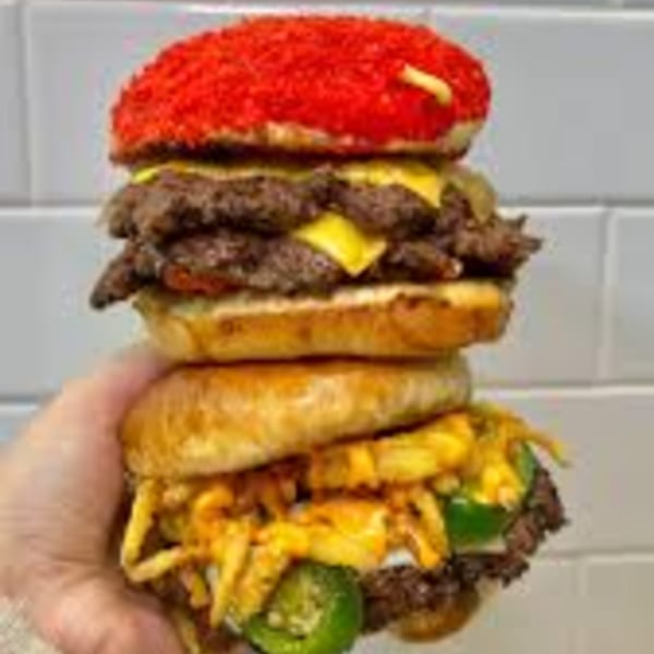 A hand holds two stacked Rodeo Burgers: one with a red bun, the other with jalapeños and fries.