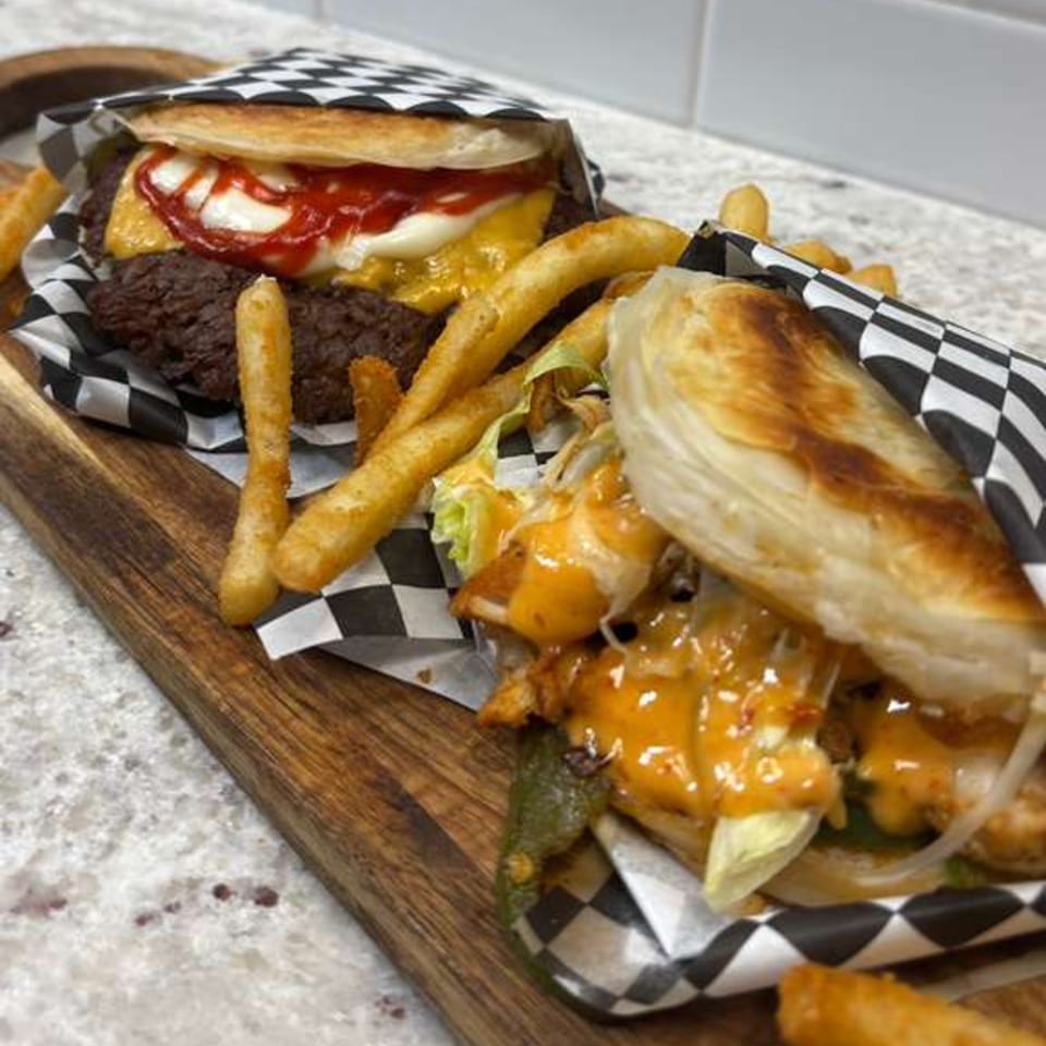 Two Rodeo Burgers and Chicken sandwiches with fries, wrapped in checkered paper on a wooden board.