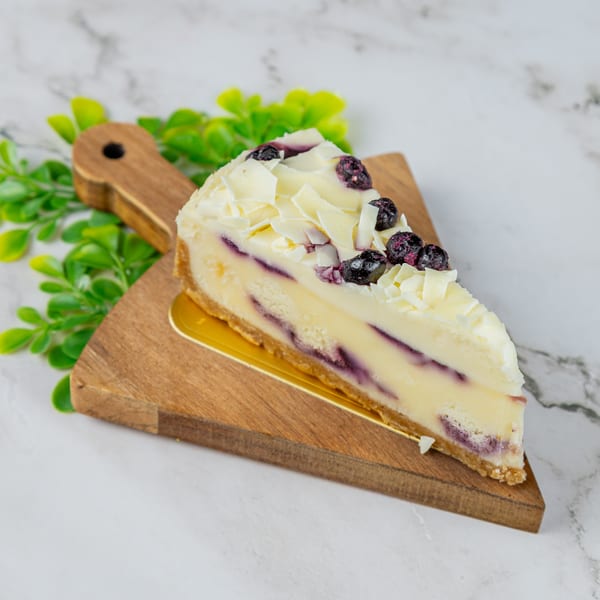 A slice of blueberry cheesecake with white chocolate shavings on a wooden board.